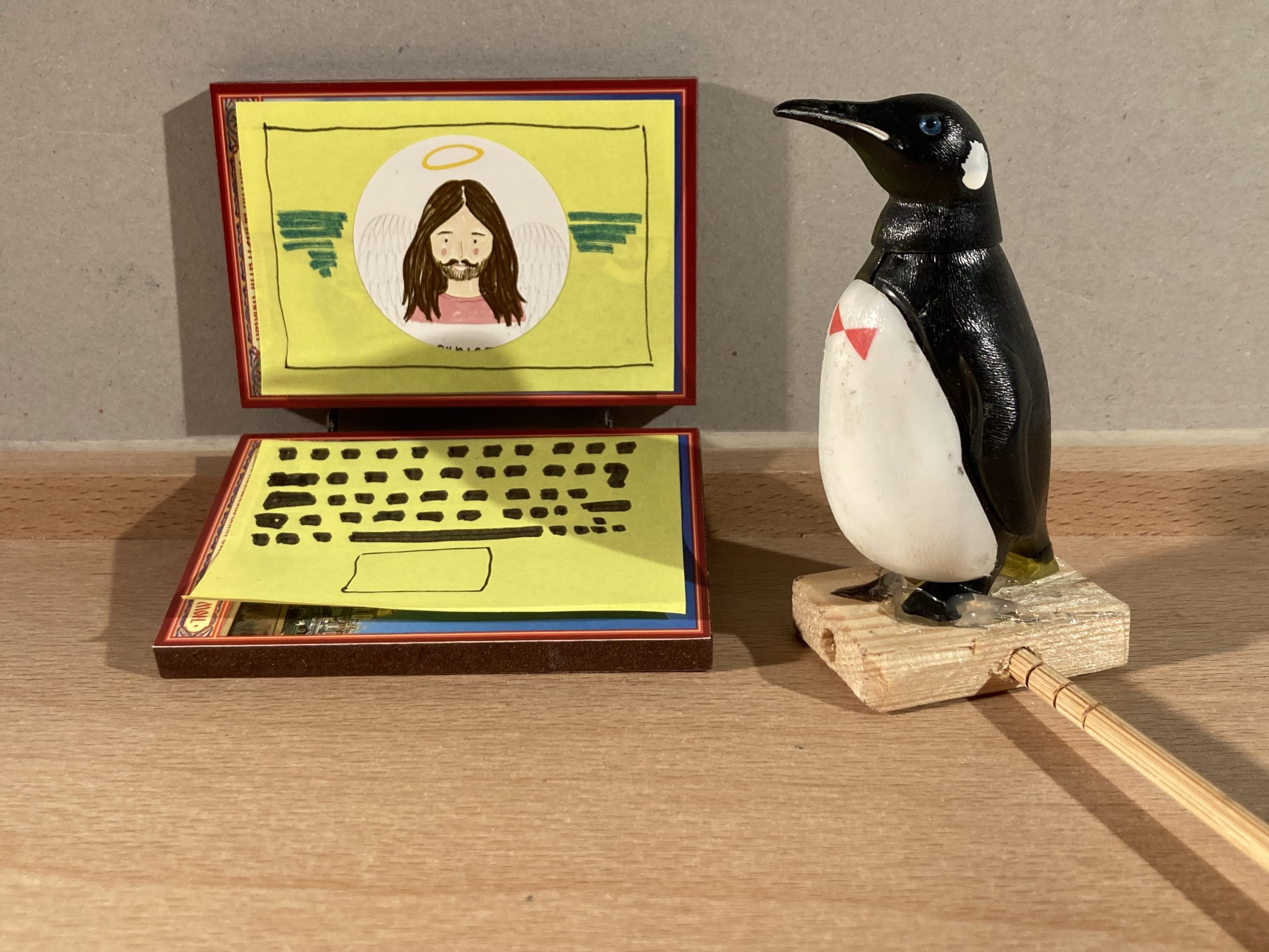Rechts im Bild ist ein Pinguin. Er schaut sich den Laptop an. Auf dem Bildschirm des Laptops ist ein Mann mit langen Haaren zu sehen. Er lächelt.